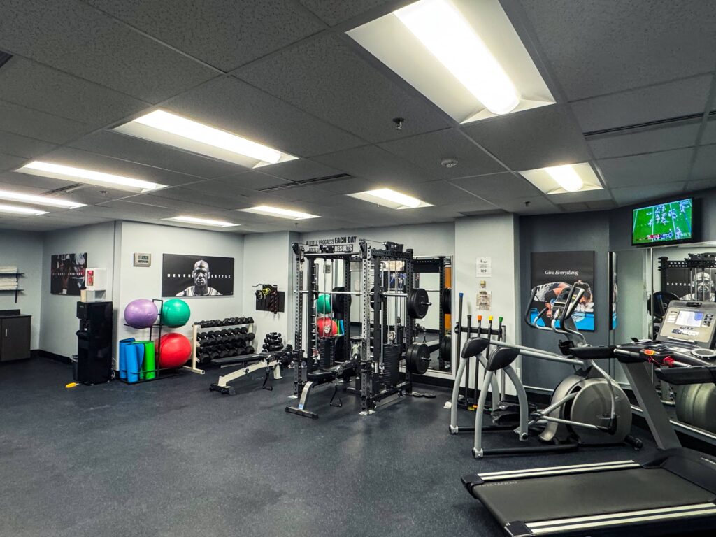 The fitness center inside the HeinOnline Business & Technology Center in Getzville, New York, featuring treadmills, stationary bikes, a cable weight machine, dumbbells, resistance balls, yoga mats, and motivational posters on the walls. The room is well-lit with overhead fluorescent lights, and a TV in the corner displays a sports game.