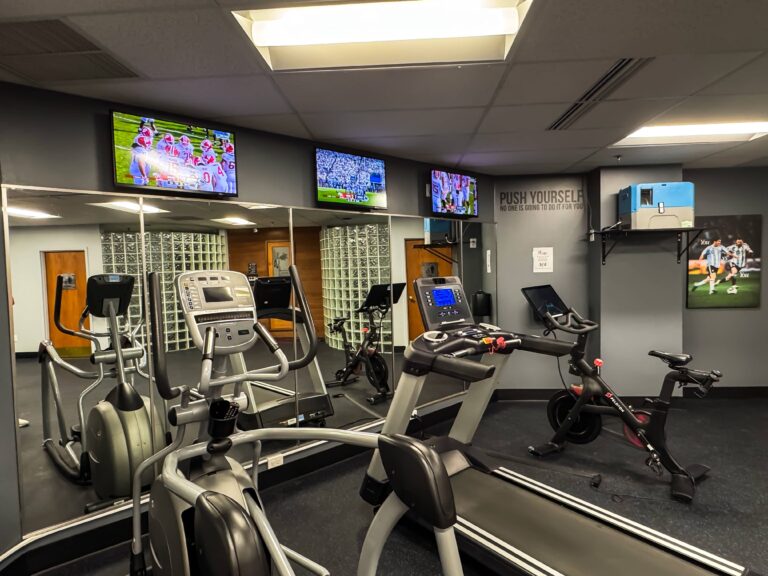 Cardio area inside the HeinOnline Business & Technology Center fitness center in Getzville, New York, featuring treadmills, an elliptical machine, and a stationary bike facing mirrored walls and mounted TVs displaying sports games. Motivational wall art and a “Push Yourself” quote decorate the space.