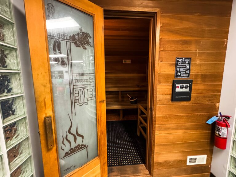 A wooden sauna room inside the HeinOnline Business & Technology Center fitness center in Getzville, New York. The partially open glass door features an etched sauna design, revealing wooden benches and a bucket inside. A fire extinguisher and control panel are mounted on the adjacent wall.