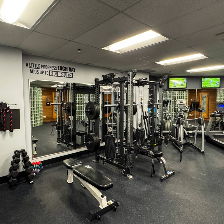 Weight training area inside the HeinOnline Business & Technology Center fitness center in Getzville, New York, featuring a cable crossover and squat rack, free weights, and a bench positioned near mirrored walls. Motivational text on the wall reads, “A little progress each day adds up to big results,” with cardio equipment and TVs visible in the background.
