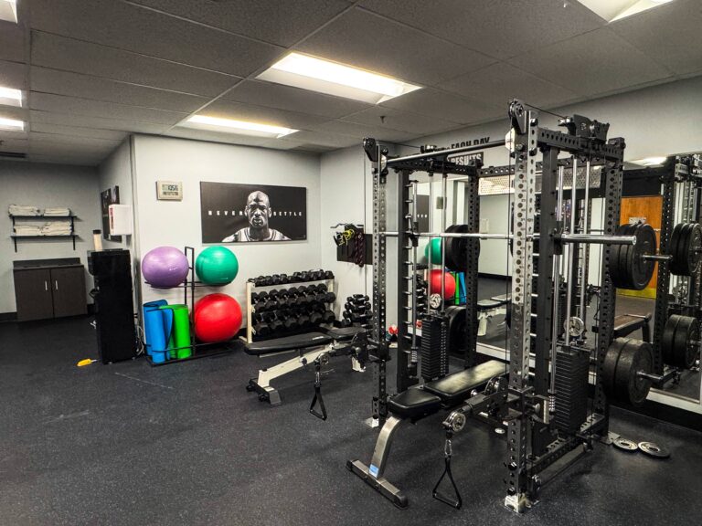 Strength training area inside the HeinOnline Business & Technology Center fitness center in Getzville, New York, featuring a power rack with weight plates, dumbbells, benches, stability balls, and yoga mats. The walls display motivational posters, and clean towels are stacked on shelves nearby.